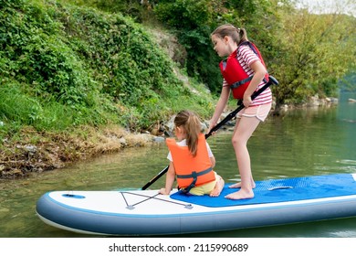 Girls Padding On Stand Up Paddle Boarding On Lake District. Children In Swim Life Vest Learning Paddleboarding On SUP Board. Family Active Leisure And Local Getaway With Kids Concept