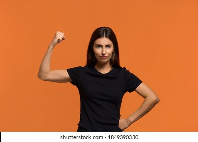 Girls power. Serious caucasian woman in black t-shirt raising hand while showing muscle, trying to look dangerous while. Ready to revenge everyone for bad things. Isolated over an orange background.