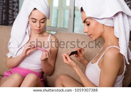 Girls are preparing for a party or a walk. Two young beautiful smiling women with towels on their heads sitting at home on the couch talking, doing a manicure and makeup before the party.
