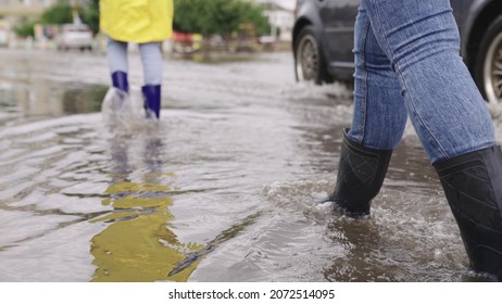 girls in raincoats and rubber boots walk along road flooded with torrential rains, their feet walk through puddles city, splashing water to the sides, the flood is on street, car is driving on water