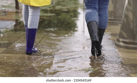 Girls In Raincoats And Rubber Boots Walk Along Road Flooded With Torrential Rains, Their Feet Walk Through Puddles City, Splashing Water To The Sides, The Flood Is On Street, Car Is Driving On Water