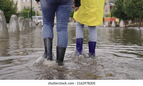 girls in raincoats and rubber boots walk along road flooded with torrential rains, their feet walk through puddles city, splashing water to the sides, the flood is on street, car is driving on water. - Shutterstock ID 2162978205