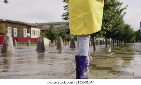 Girls In Raincoats And Rubber Boots Walk Along Road Flooded With Torrential Rains, Their Feet Walk Through Puddles City, Splashing Water To The Sides, The Flood Is On Street, Car Is Driving On Water