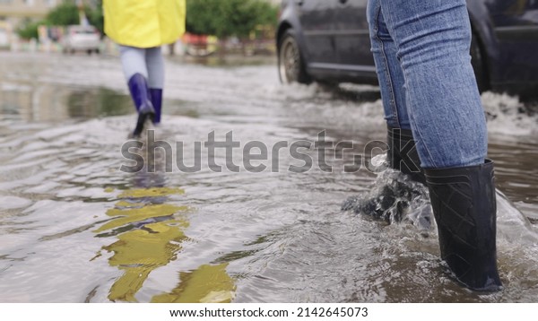 girls in raincoats and rubber boots walk along\
road flooded with torrential rains, their feet walk through puddles\
city, splashing water to the sides, the flood is on street, car is\
driving on water.