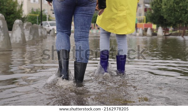 girls in raincoats and rubber boots walk along\
road flooded with torrential rains, their feet walk through puddles\
city, splashing water to the sides, the flood is on street, car is\
driving on water.