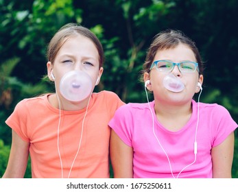 Girls Sharing Headphones To Listen To Music. The Sisters Chew Gum And Make Balloons Out Of It. Happy Friends Spending Time Together