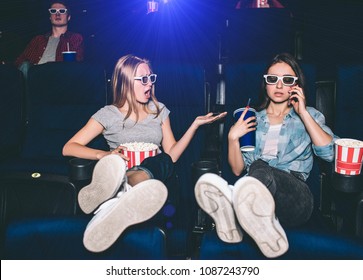 Girls Are Sitting In Chairs In Cinema Hall. Brunette Is Talking On The Phone While Her Friend Is Making Remark To Her. Girl On Th Left Is Upset And Irritated.