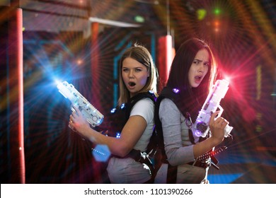 Girls standing back to back in colorful laser beams, holding guns during lasertag game in labyrinth