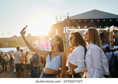 Girls in summer attire enjoy sunset at coastal music festival, stage lights glow in background. Youth share moments, dance to live performances, sandy beach venue vibrates with fest energy. - Powered by Shutterstock