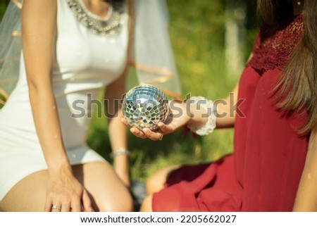 Girls in summer in dresses sit on grass. Girl holds ball in her hand. Mirror ball in palm of your hand. Details of outdoor recreation.