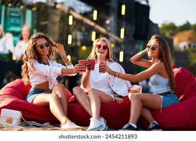 Girls in summer outfits sit on red bean bag chairs, clinking cups at beach music festival. Laugh, enjoying fest vibes, relax with drinks, sunset, ocean backdrop. Casual celebration cheerful friends. - Powered by Shutterstock