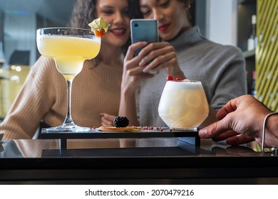 girls taking cocktails pictures with smartphone - food blogger influencers shooting at restaurant - bartender serving vodka sour and margarita cups - focus on glasses