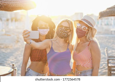 Girls taking a selfie with face mask outside for coronavirus protection - Teenagers in vacation at a beach enjoying the sunset - Multiracial youth having fun together - Young people using smartphone - Powered by Shutterstock