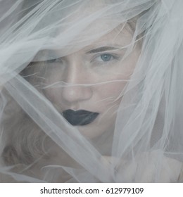girl's or women's face behind a veil.  Portrait of a beautiful blue-eyed girl with black lips behind a white veil.  Black lips. tender portrait. Tulle.bewitching glance.Penetrating look