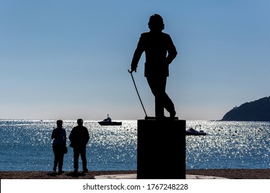 Cadaqués, Girona, Spain; 10/21/2017 Statue Of The Painter Salvador Dalí Backlit By The Morning Sun, And Silhouettes Of Two People Walking And Boats In The Glowing Sea