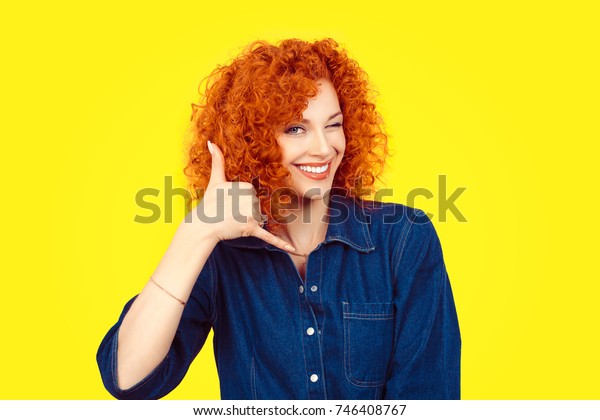 Give me a call. Closeup portrait young red\
head curly single woman excited happy student winking with eye\
making showing call me gesture sign with hand shaped like phone\
isolated yellow\
background.