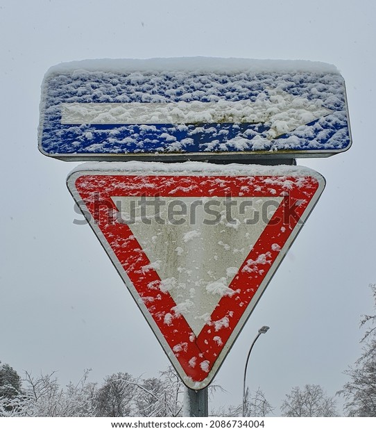Give way and one-way street traffic signs covered in\
snowflakes and road mud