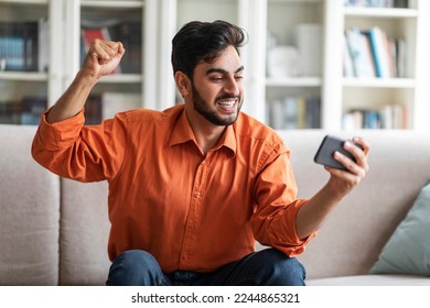 Giveaway, lottery, bet on Internet. Emotional happy handsome millennial arabic guy sitting on couch at home, gaming online, holding smartphone and gesturing raising fist up, copy space