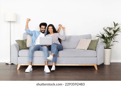 Giveaway, lottery, cashback. Emotional happy beautiful indian couple sitting on couch at home, gesturing and exclaiming, celebrating success, gambling on Internet, using laptop, copy space - Powered by Shutterstock