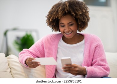 Giveaway, lottery, online bet. Emotional pretty young african woman in casual outfit resting alone on couch at home, using brand new smartphone, checking her lottery ticket online, copy space - Shutterstock ID 2253955191