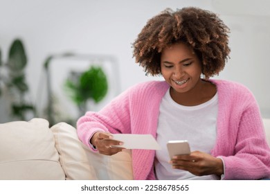 Giveaway, lottery, online bet. Excited pretty african american woman in casual outfit resting alone on couch at home, using brand new smartphone, checking her lottery ticket online, copy space