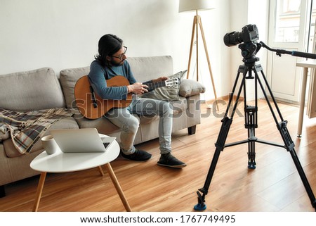 Giving guitar online lesson. Young man blogger or music teacher sitting on sofa with a guitar, recording video tutorial at home. Focus on a man. Stay home, quarantine. E-learning. Online education