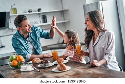 Giving High Five To Daddy While Eating In The Kitchen. Happy Family Are Enjoying Spending Time Together While Standing On Light Modern Kitchen.