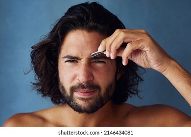 Giving His Brows Some Defined Shape. Studio Portrait Of A Handsome Young Man Plucking His Eyebrows Against A Blue Background.