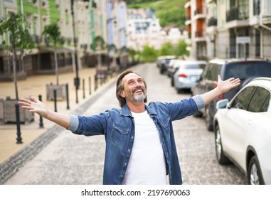 Giving Thanks To God Mature Man Standing Outdoors With Hands Spread Wide Open Wearing Jeans Shirt With White T-shirt Under. Mid Aged Man Glad That Life Is Beautiful Standing In Old Town Street. 