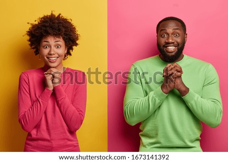 Glad Afro American girlfriend and boyfriend keep hands together in praying gesture, anticipate important results, stand next to each other against two colored background, smile broadly, feel happy