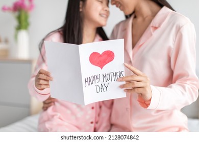 Glad Asian Teen Chinese Girl And Millennial Female In Pajamas Hold Postcard In Bedroom Interior, Cropped. Mothers Day Celebration At Home Due Covid-19 Outbreak. Love, Mom And Daughter Relationship