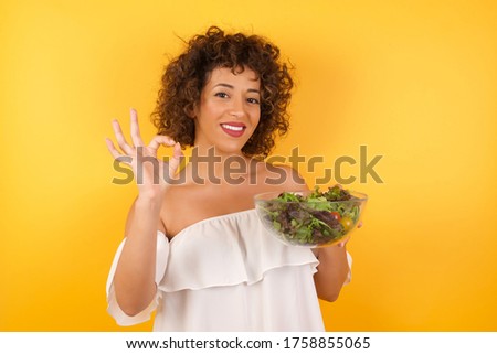 Glad attractive woman shows ok sign with both hands as expresses approval, has cheerful expression. Photo of beautiful female has appealing appearance, being optimistic. Standing against yellow wall.