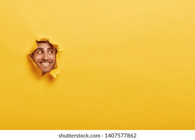 Glad Caucasian man with toothy smile, has bristle, looks positively aside, shows face in paper hole, isolated over yellow background with blank space. Positive emotions. Man peeks through ripped paper