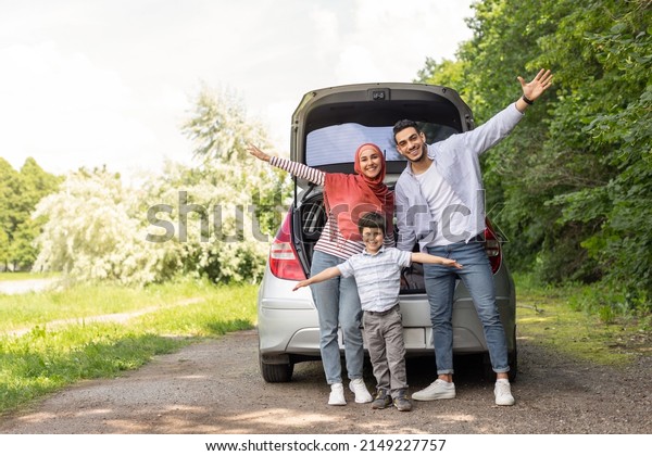Glad cheerful emotional arab male, female in hijab\
and little boy waving their hands near car trunk, enjoy journey\
together outdoor in summer. Family trip, vacation and travel at\
auto, free space