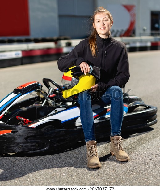 Glad cheerful positive smiling\
woman with helmet sitting in car for karting in sport\
club