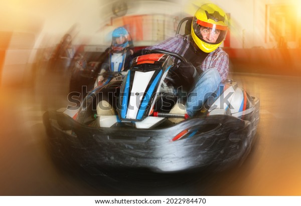Glad cheerful smiling male\
in helmet and other people driving cars for karting in sport club\
indoor