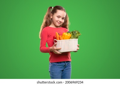 Glad Child Carrying Basket Of Healthy Vegetables And Looking At Camera Against Green Background