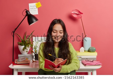 Glad concentrated woman focused in notepad, writes down ideas for essay or research work, makes up review, poses against workplace with desk lamp, textbooks, calla lilies in vase, pink background