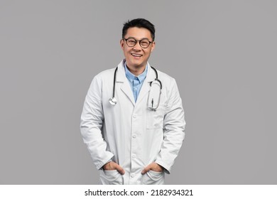 Glad Confident Attractive Adult Japanese Doctor In White Coat, Glasses With Stethoscope Look At Camera Isolated On Gray Background, Studio. Health Care, Medical Business, Consultation And Treatment