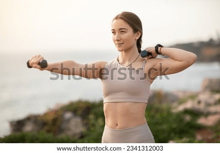 Glad confident strong millennial caucasian woman athlete in sportswear doing arm exercises with dumbbells, enjoy workout, body care alone at sea beach. Sports, fitness outdoor