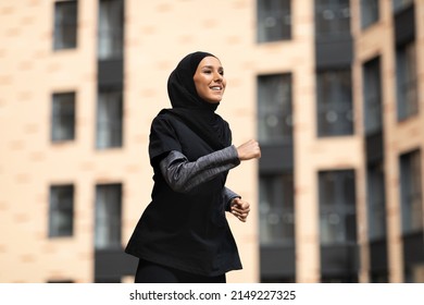 Glad confident young muslim woman in black hijab and sportswear runs outdoors, on building background, free space. Vitality, urban workout season, jogging, sports in city, health care and weight loss - Powered by Shutterstock