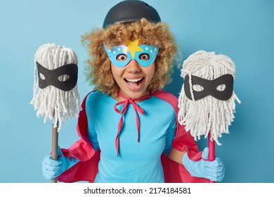 Glad Curly Woman Pretends Having Super Power And Being Cleaning Hero Wears Helmet Mask Cloak And Rubber Gloves Holds Two Mops Ready To Bring Your House In Order Isolated Over Blue Background