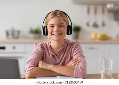 Glad Cute European Teen Blonde Girl In Headphones Study At Home At Table With Glass Of Water And Laptop In Kitchen Interior. Covid-19 Quarantine, Remote Schooling, Knowledge, Education And Health Care