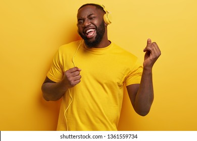 Glad dark skinned plump man dances, makes movements to music, has modern stereo headphones, smiles positively, being in high spirit. Everything in yellow colour. Carefree guy listens lively music