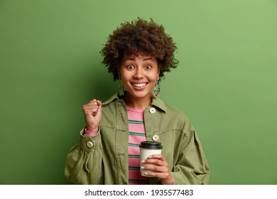 Glad dark skinned young woman clenches fist celebrates positive news smiles happily drinks takeaway coffee wears stylish clothes isolated over green background. People emotions lifestyle concept