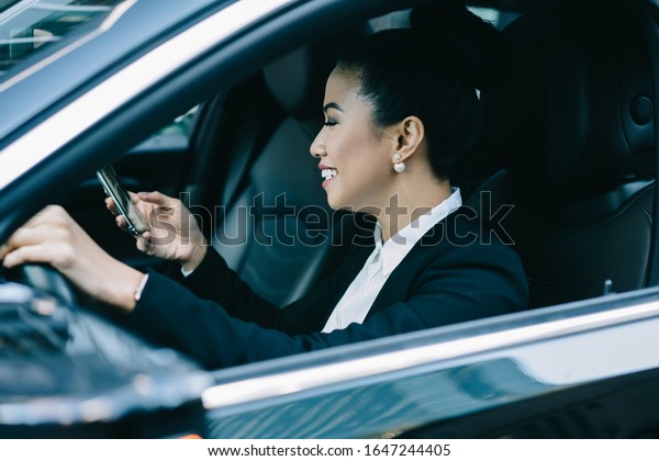 Glad ethnic female in formal clothes steering\
automobile driving modern car chatting on mobile messaging on phone\
sitting at wheel