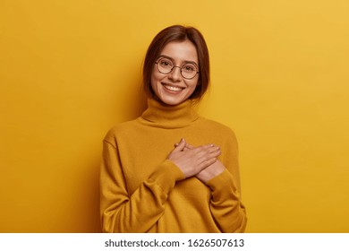 Glad European Girl Feels Pleased And Grateful, Being Thankful For Awesome Gift, Presses Hands On Chest, Has Pleasant Tender Smile, Tilts Head, Wears Yellow Turtleneck. What Pleasure To Meet You