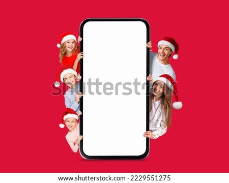 Glad european millennial family with kids in Santa Claus hats peeking out from big smartphone with empty screen isolated on red background. Digital offer, ad and app for New Year, holiday celebrations