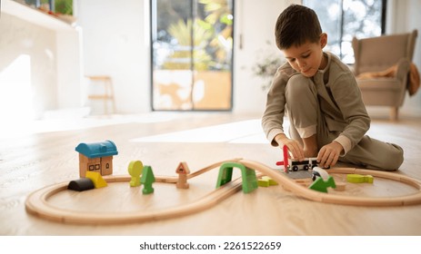 Glad european small kid in pajamas plays with toys, train, cars and wooden road, enjoy spare time, sits on floor in bright living room interior, panorama. Fun alone, fantasy at home and kindergarten