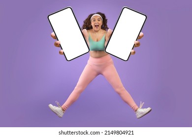 Glad Excited Young Caucasian Plus Size Woman In Sportswear Jump And Freeze In Air, Show Two Smartphones With Blank Screens, Isolated On Purple Background. Health Care, App For Training, Sale And Ad